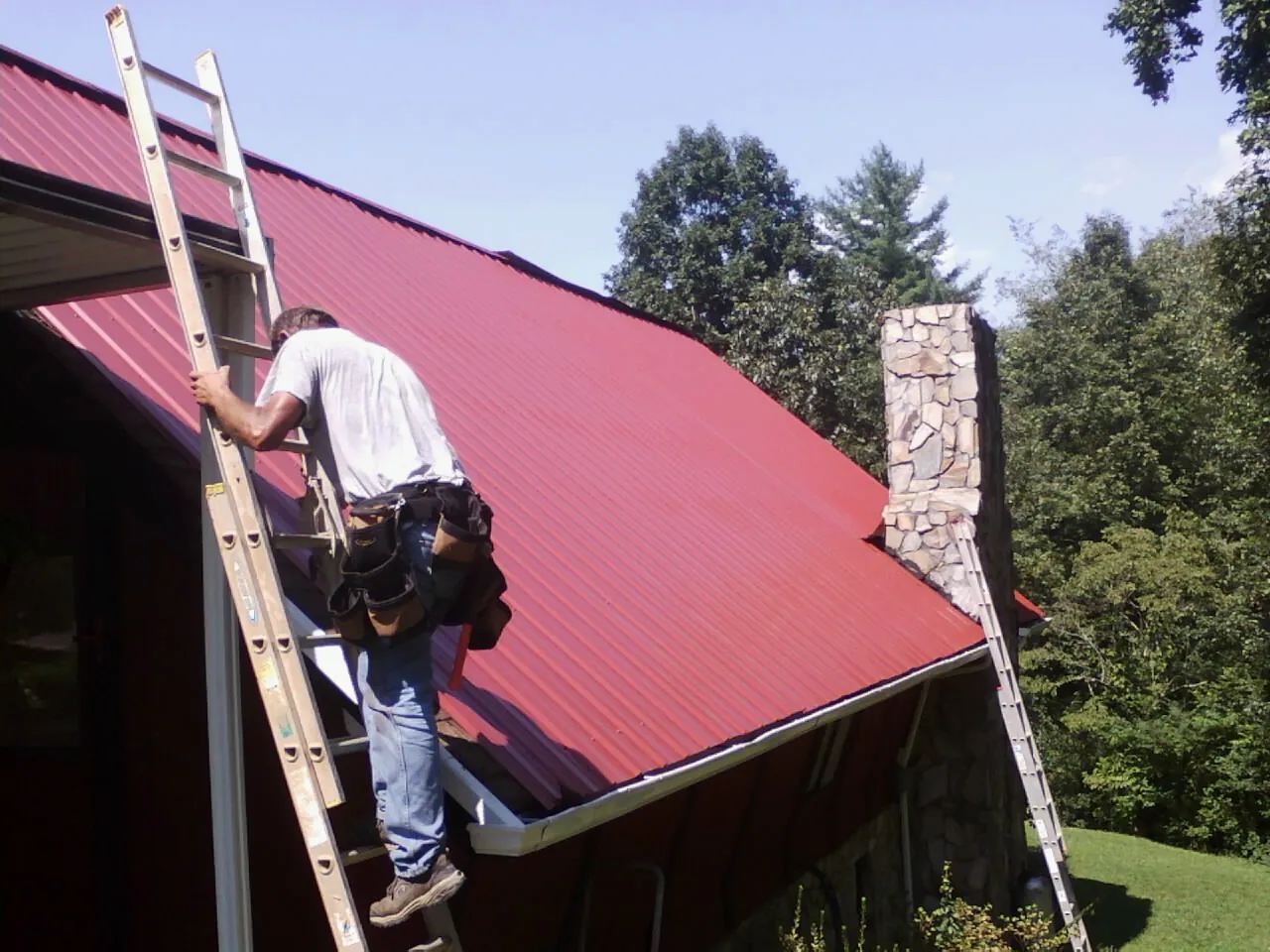 Roofer on a ladder beside a red metal roof and stone chimney — Soffit & Fascia Repair by Roofing Brothers in Rossford